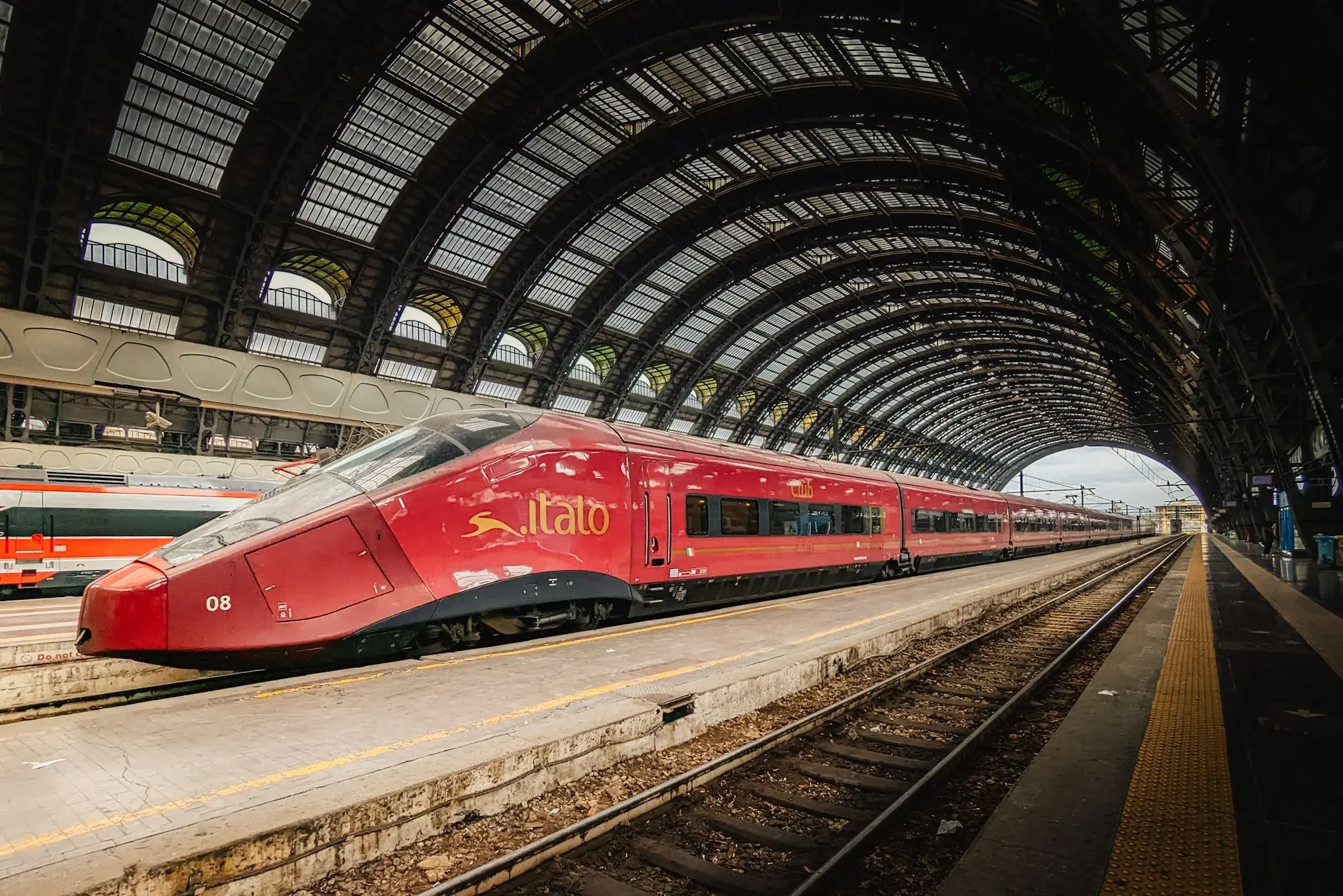 A red high-speed train under the arched iron-and-glass roof of Milano Centrale station