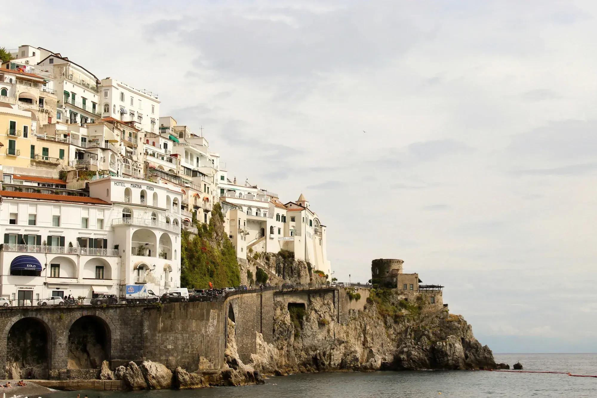 The white houses of Amalfi on a steep hillside above the sea, with the coast road running along the sea wall
