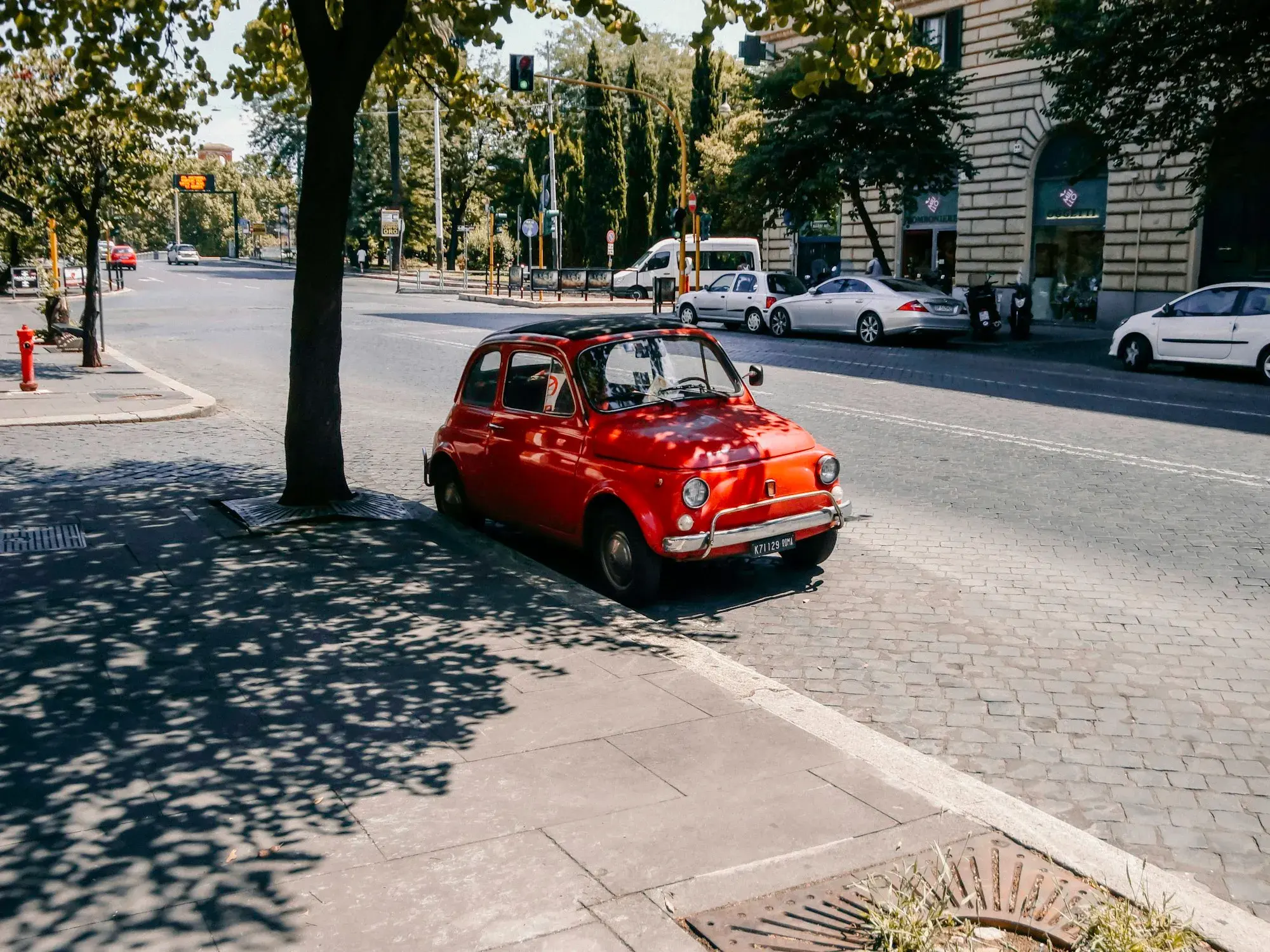 A small red Fiat 500 parked at the edge of a tree-lined street in Rome