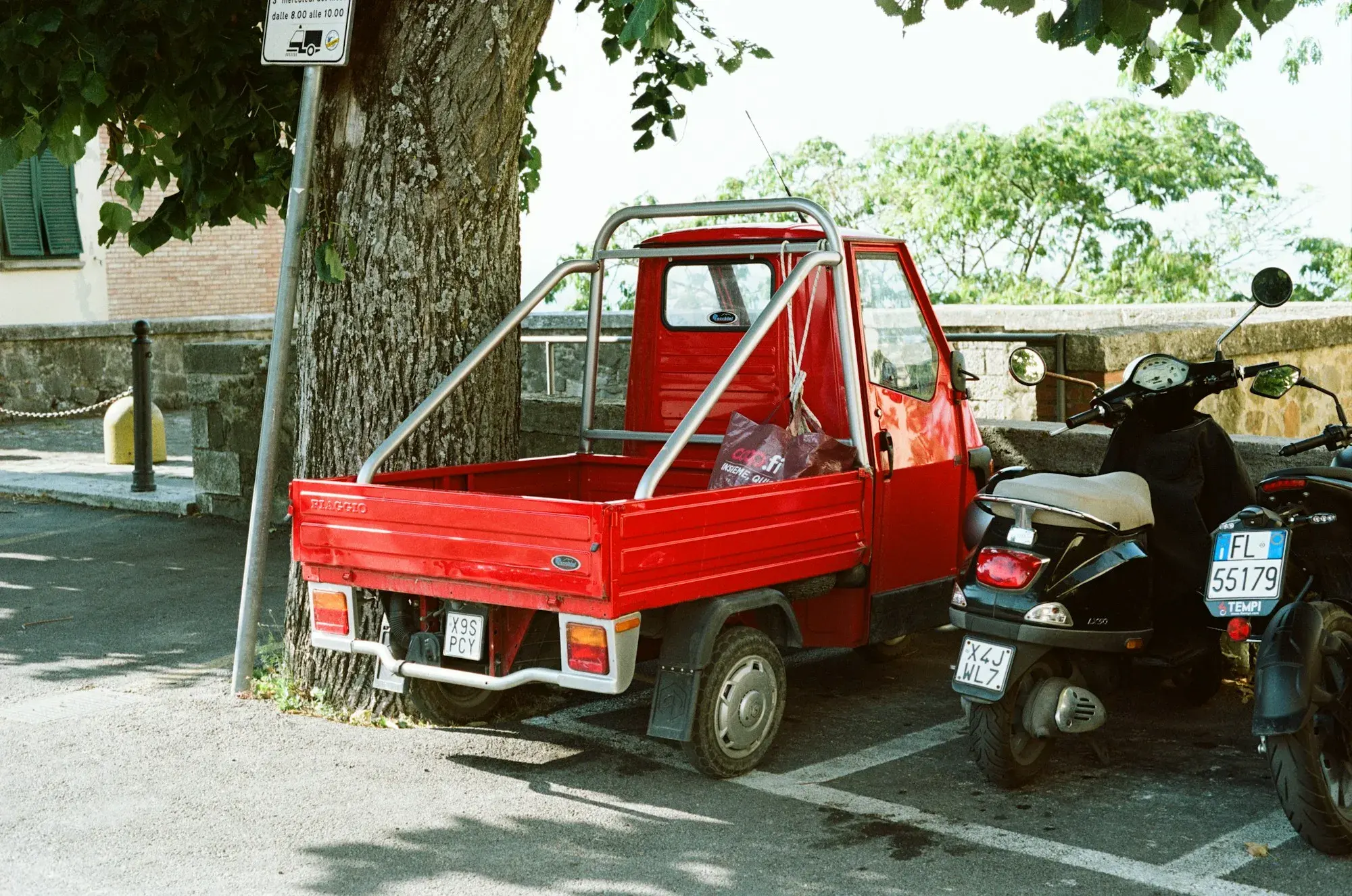 A red Piaggio Ape three-wheeler and scooters parked under a tree on a street in Montalcino