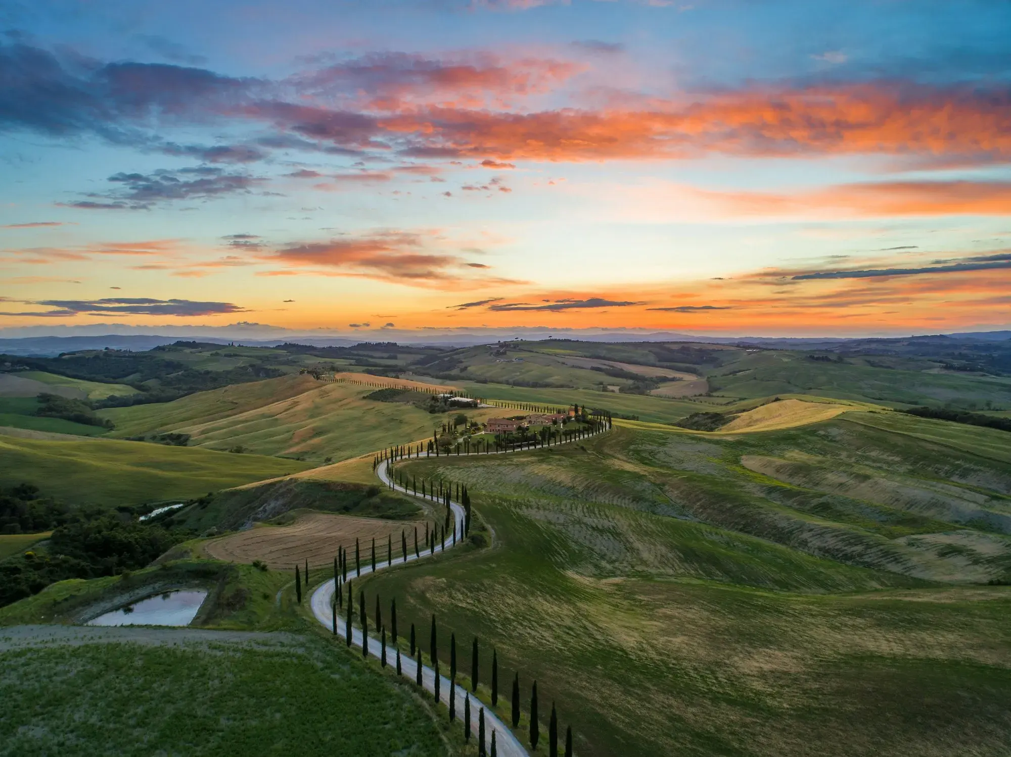 A winding road lined with cypress trees crossing rolling green hills near San Quirico d'Orcia in Tuscany