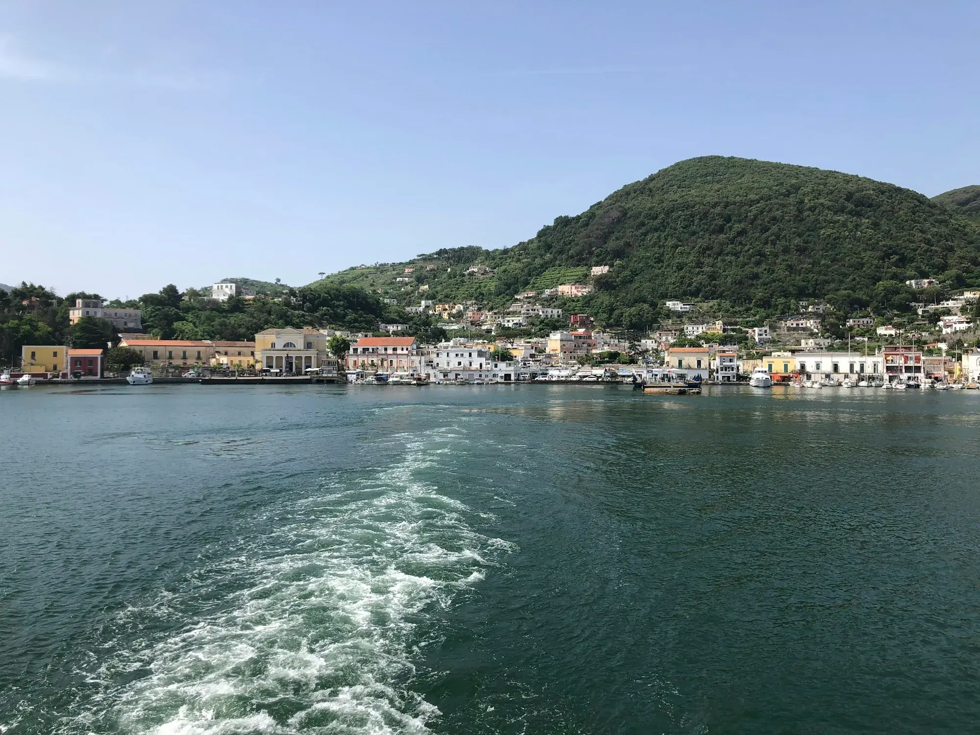 The white wake of a ferry leaving the harbour of Ischia, with the island's green hills and waterfront behind
