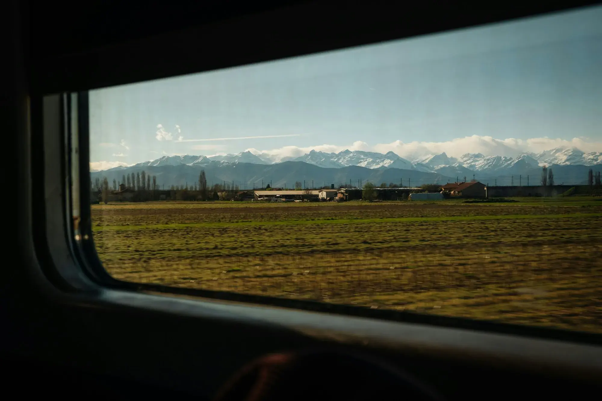 Snow-capped Alps beyond flat green fields seen through a train window near Turin