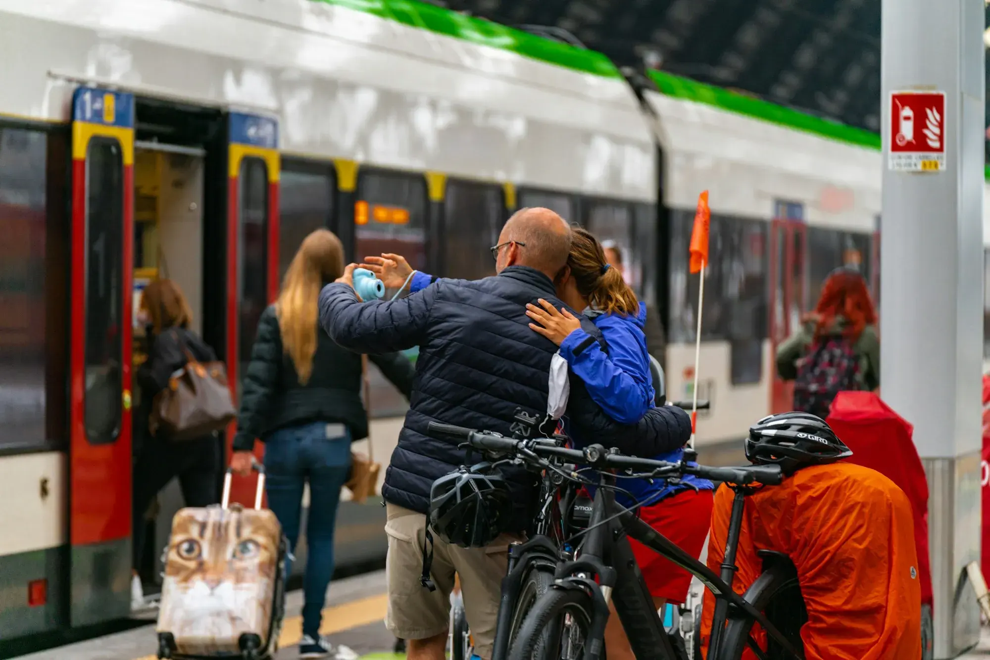 Travellers with luggage and a bicycle beside a green and white regional train on a platform at Milano Centrale