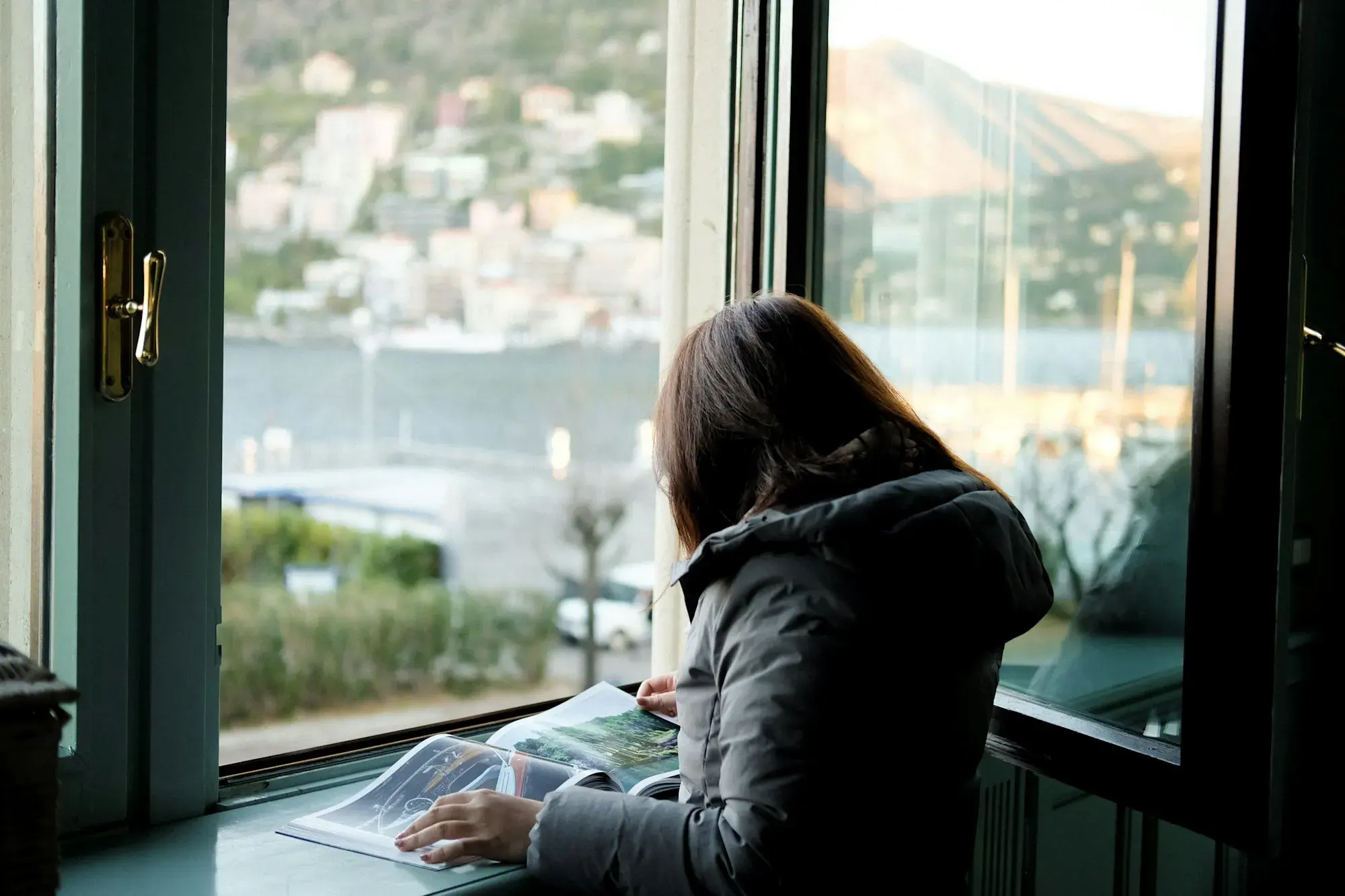 A woman reading a magazine at a window overlooking Lake Como, with houses on the far shore