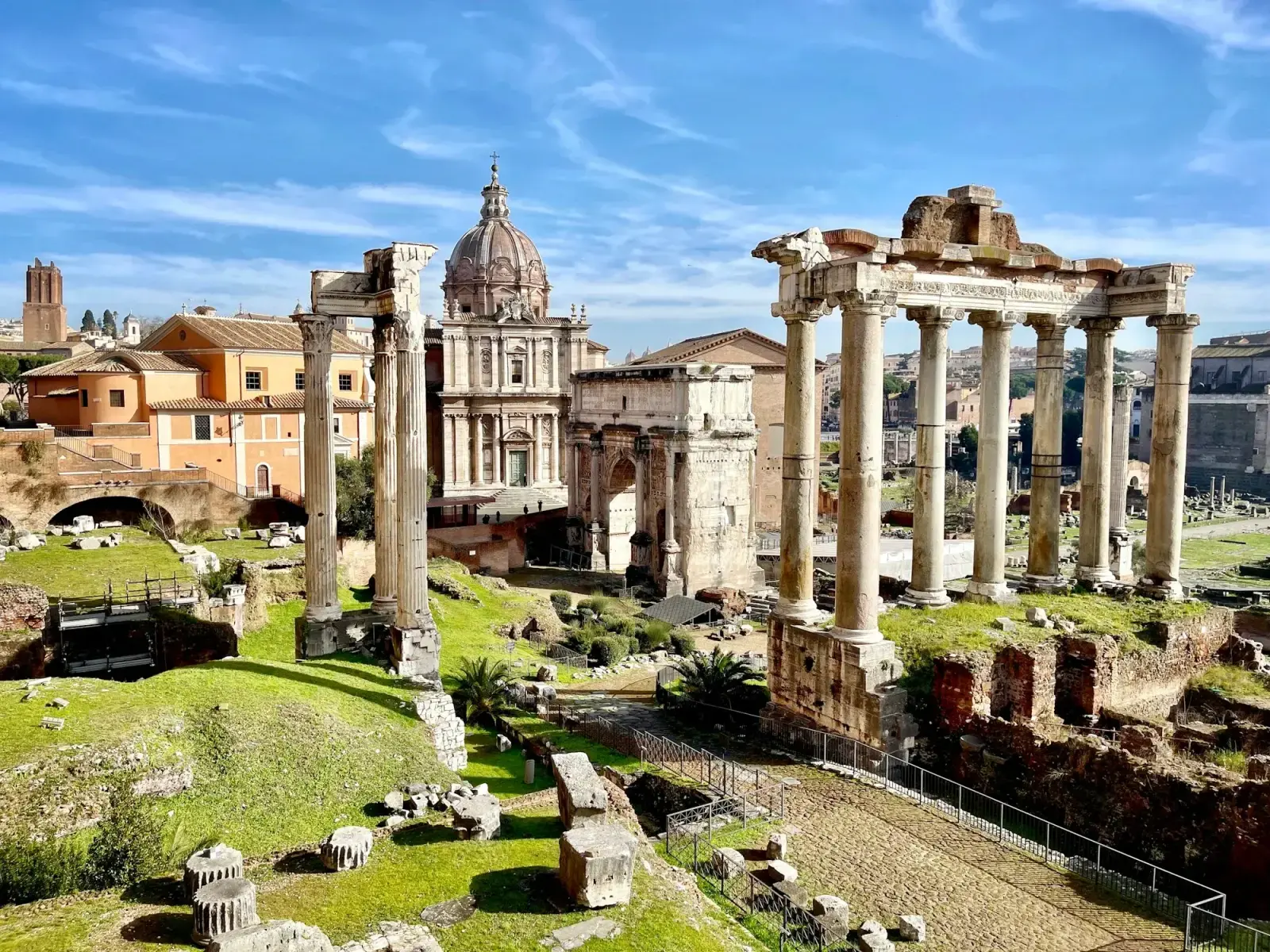 Ruins of the Roman Forum with the columns of the Temple of Saturn, a triumphal arch and church domes beyond, under a blue sky
