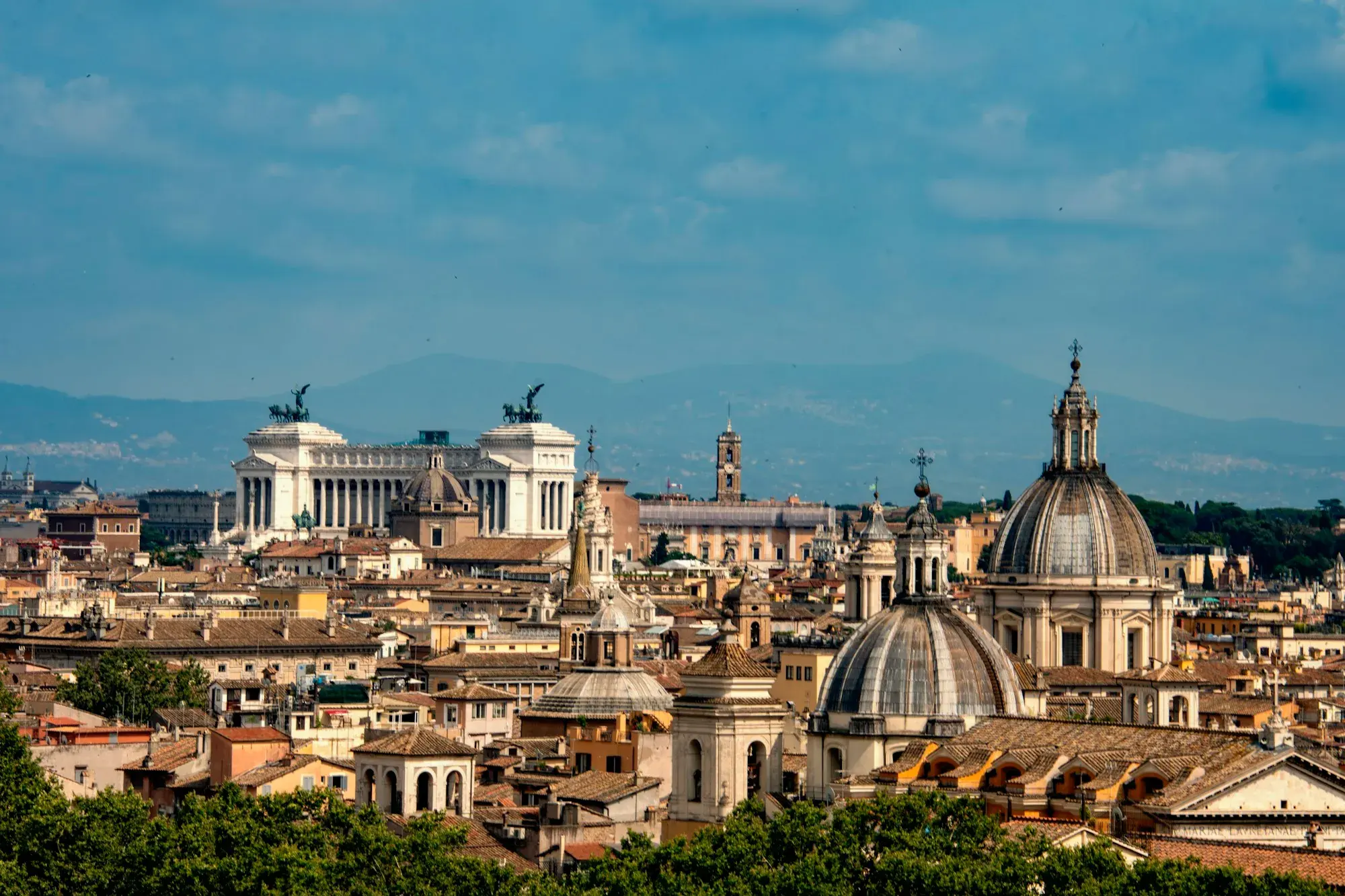 Rome's rooftops, church domes and bell towers under a blue sky, with the white Vittoriano monument on the left and hills on the horizon