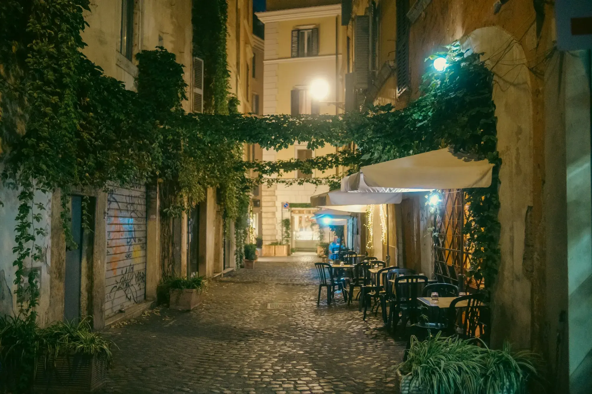 A cobbled lane in Trastevere in the evening, with ivy on the walls, lamps and restaurant tables set outside