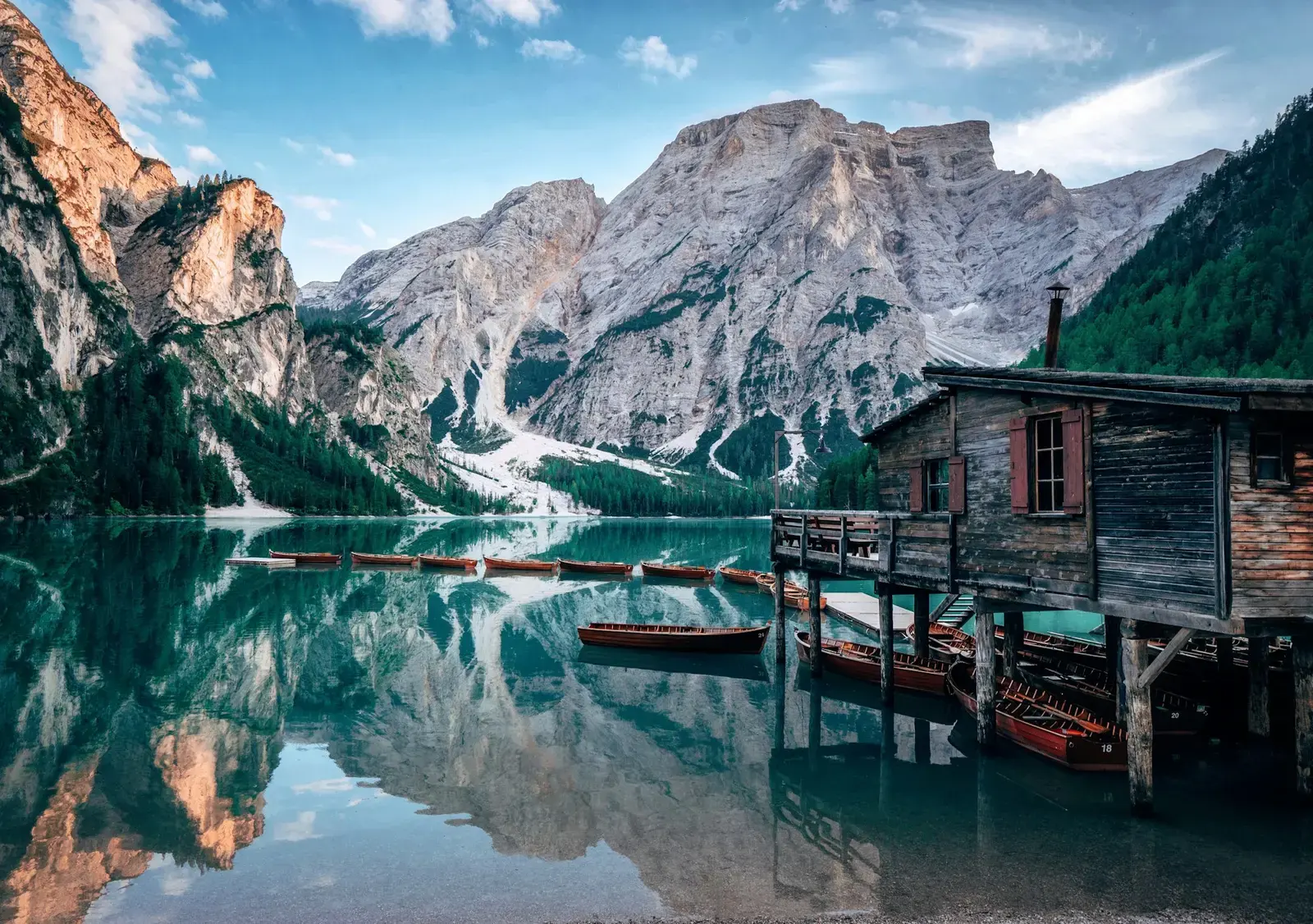 A wooden boathouse and rowing boats on the calm green water of Lago di Braies, with mountains reflected in it
