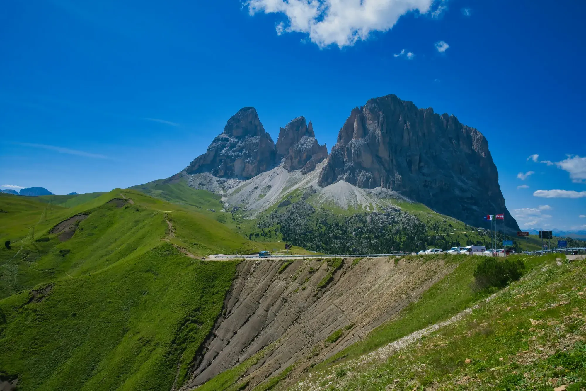 The Sassolungo and Sassopiatto peaks rising above green slopes, with a mountain road crossing the pass below