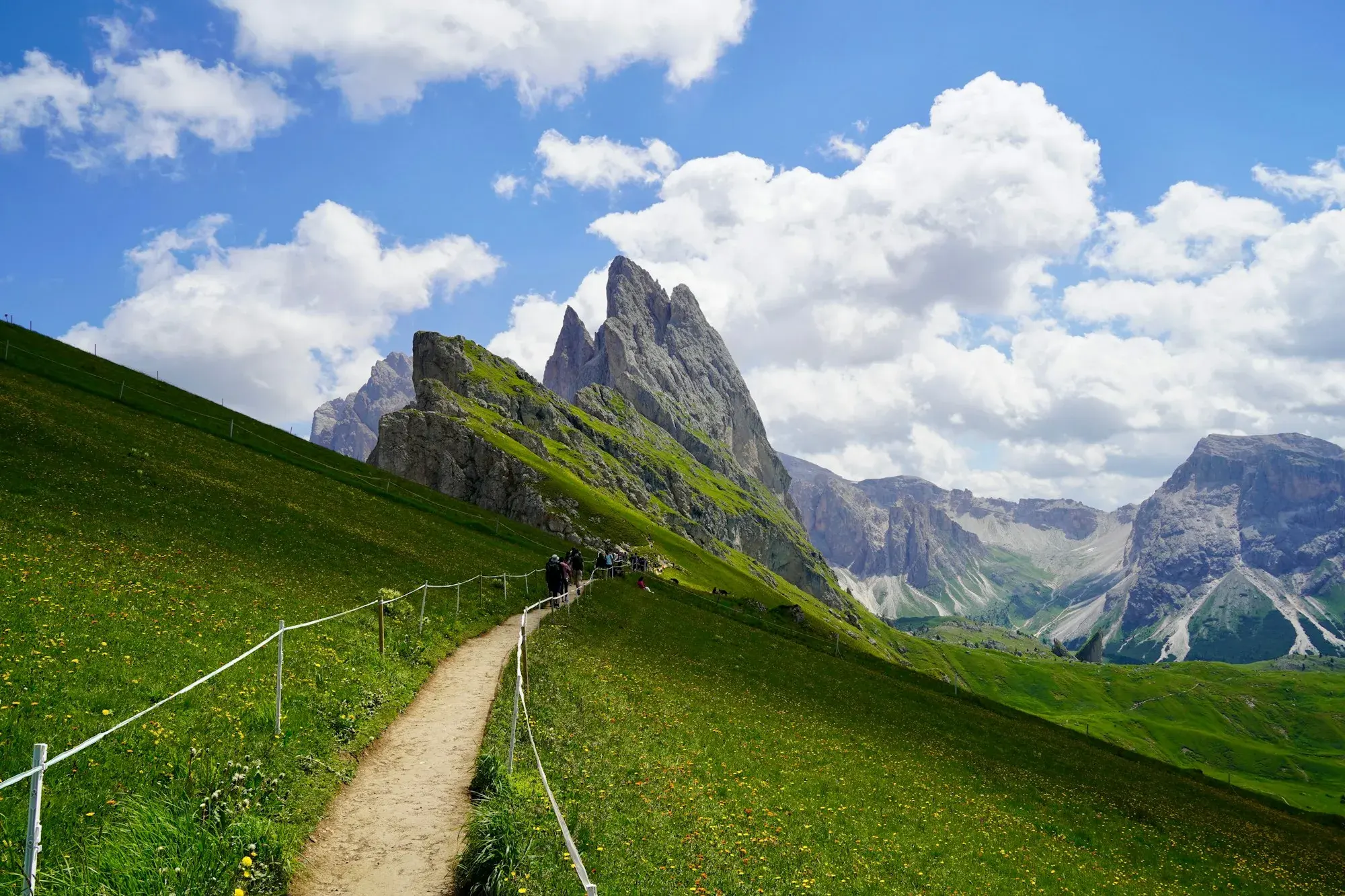 Escursionisti su un sentiero tra ripidi prati verdi verso la cresta frastagliata del Seceda, sopra Ortisei