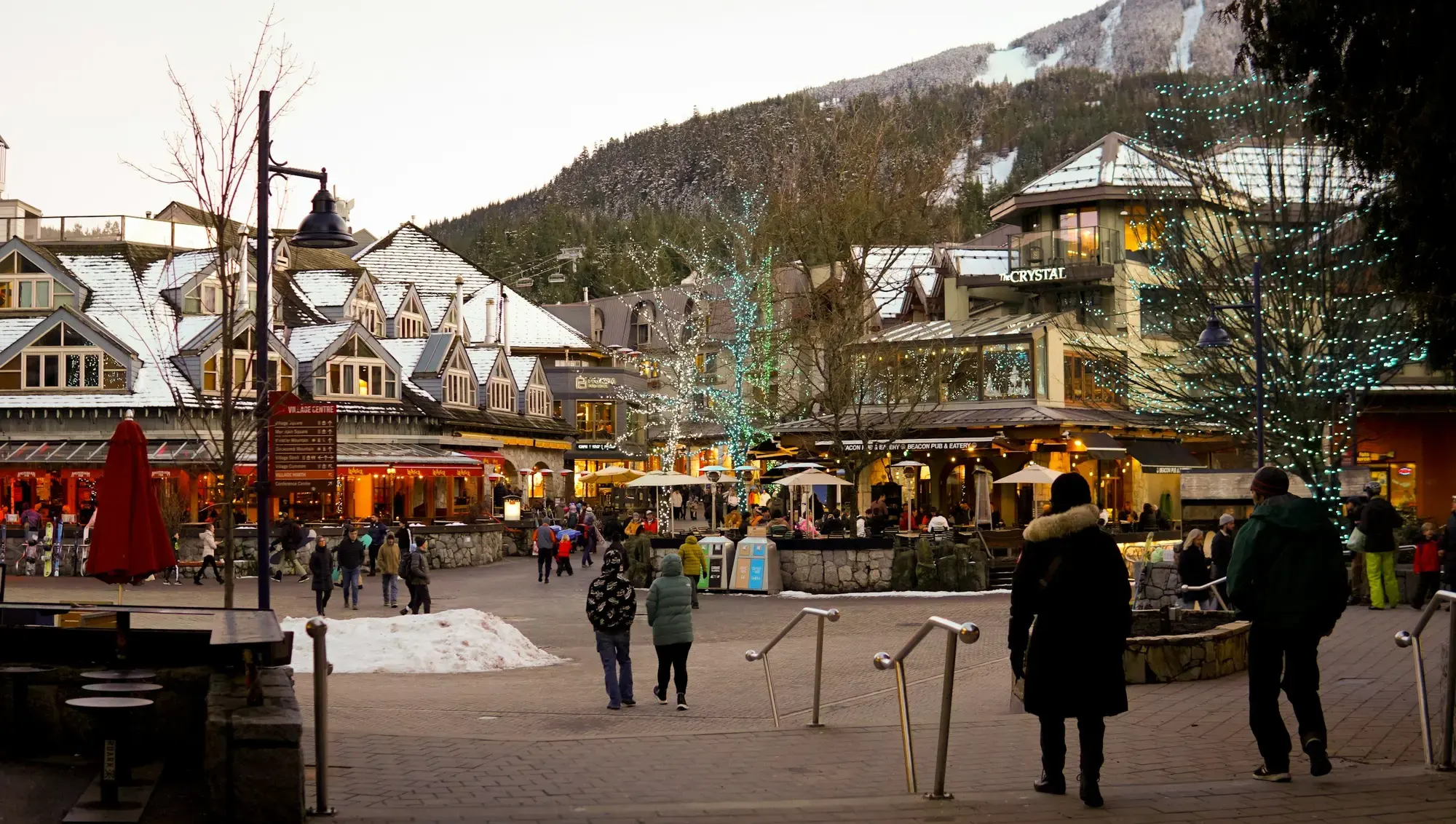 Una piazza festiva con edifici innevati e visitatori che sfogliano le bancarelle di un mercatino di Natale al tramonto