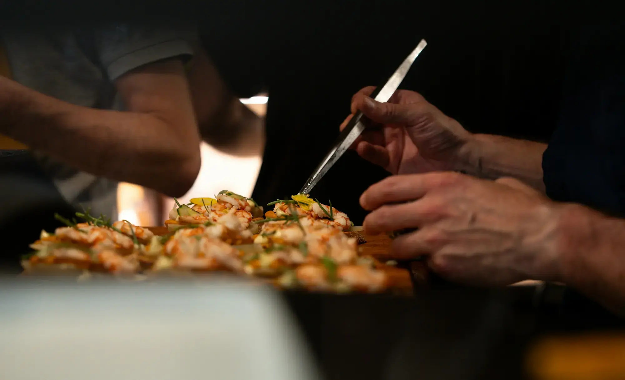 Mani che completano piccoli crostini con gamberi e altri condimenti su un bancone, nello stile dei cicchetti veneziani