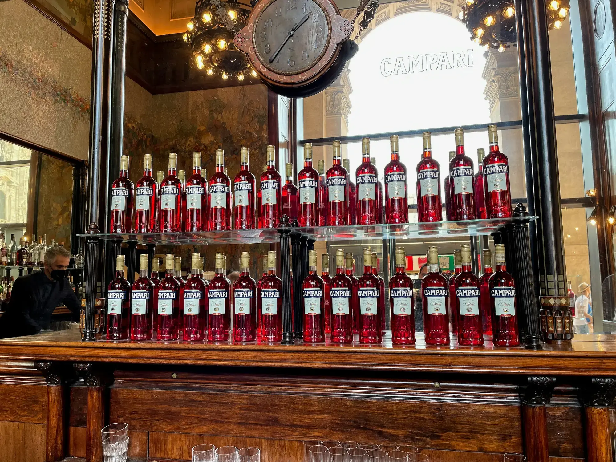 File di bottiglie di bitter rosso sugli scaffali dietro il bancone di un bar di Milano, sotto un orologio e un'insegna Campari