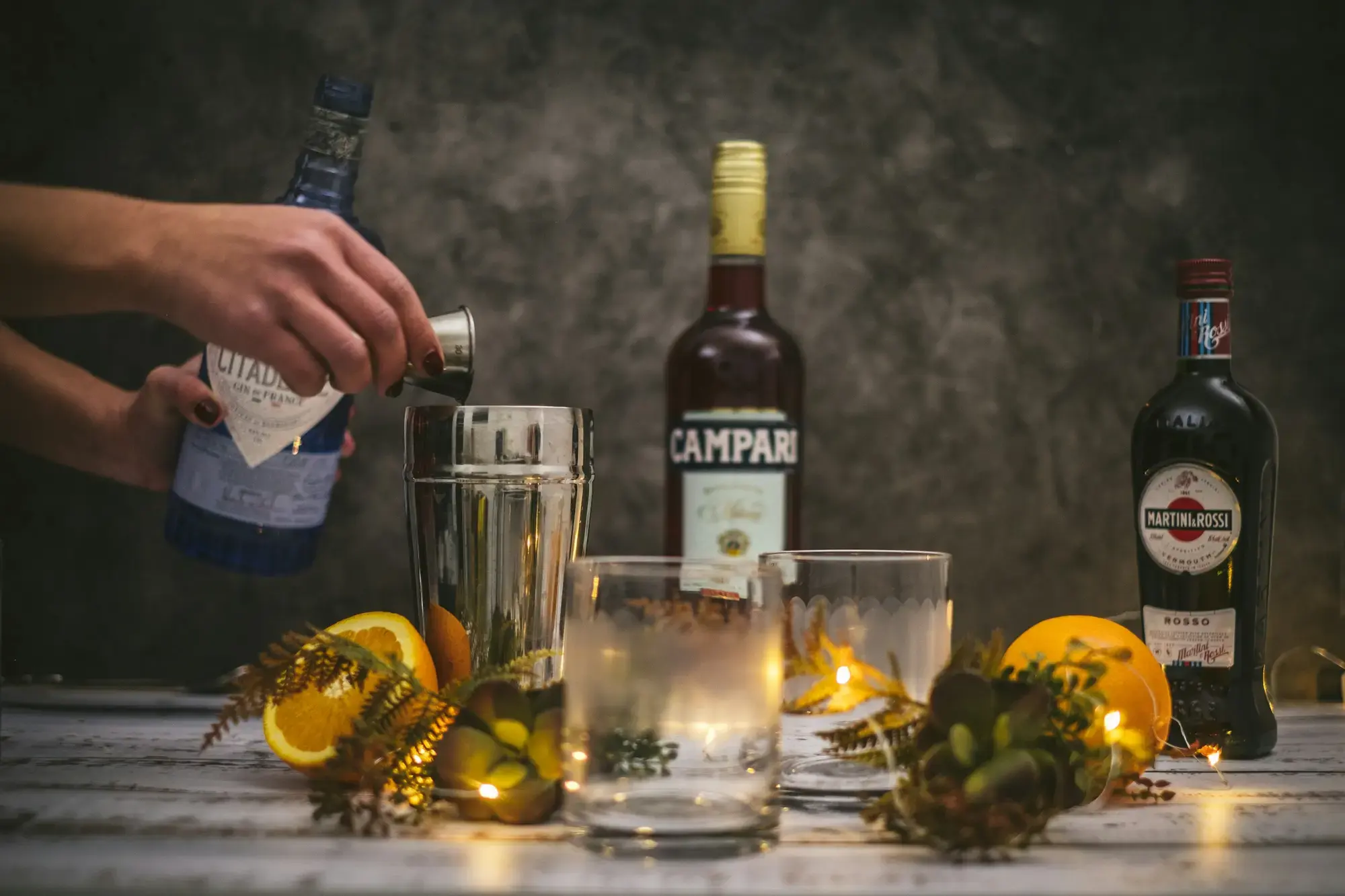 La mano di un barista versa in un mixing glass accanto a bottiglie di Campari e vermouth rosso, con bicchieri di ghiaccio e fette d'arancia