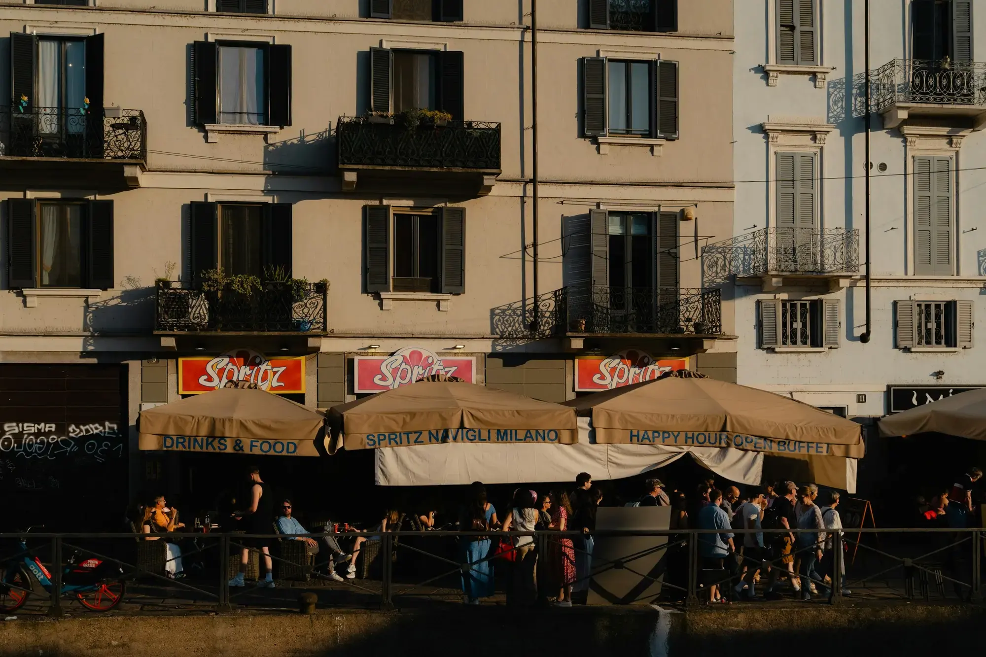 Gente ai tavoli all'aperto sotto tende che pubblicizzano spritz e happy hour lungo i Navigli a Milano, con palazzi sullo sfondo