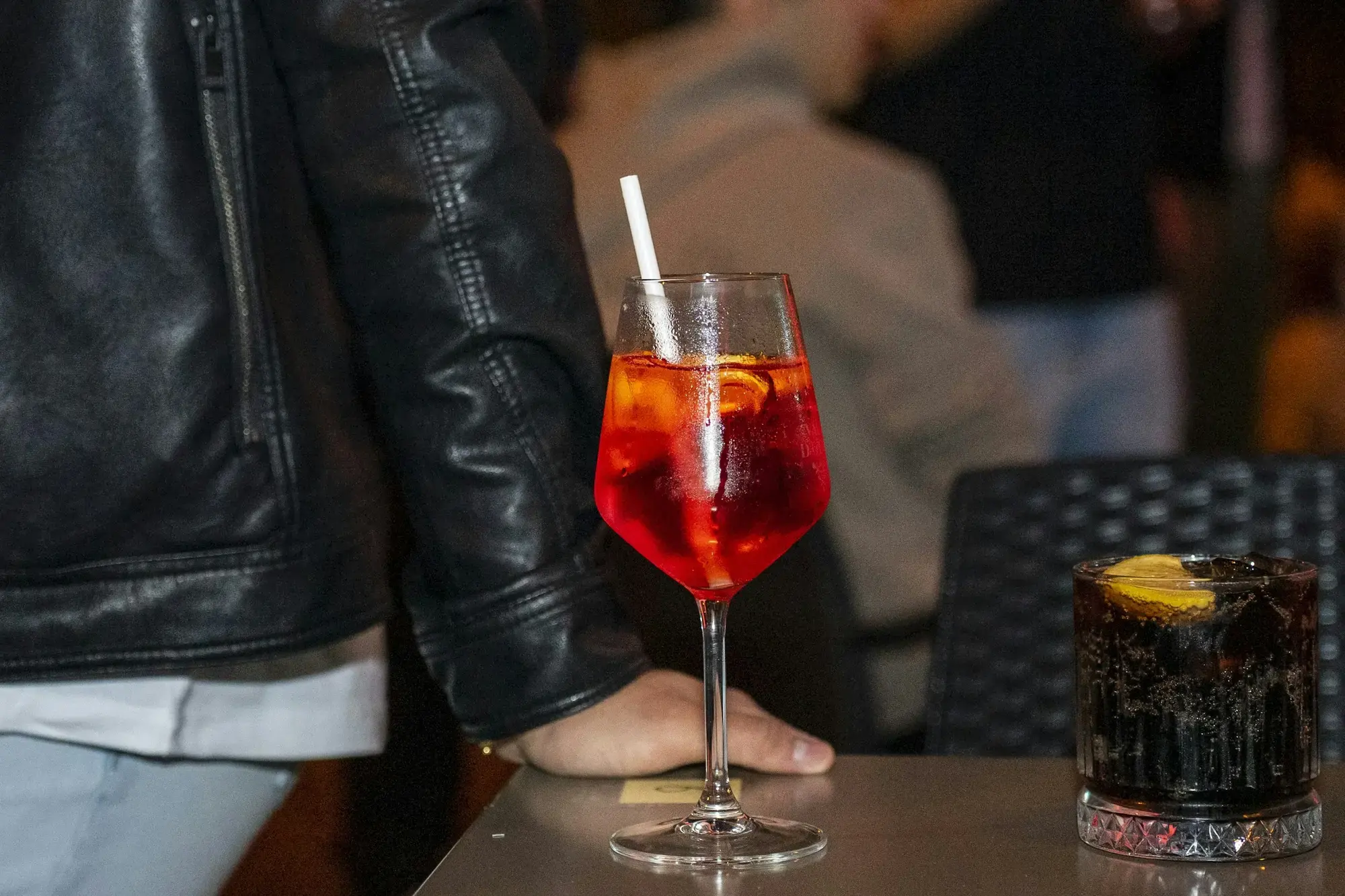 Uno spritz arancione con cannuccia e fetta d'arancia sul tavolo di un bar in Veneto, accanto a una bibita scura con ghiaccio