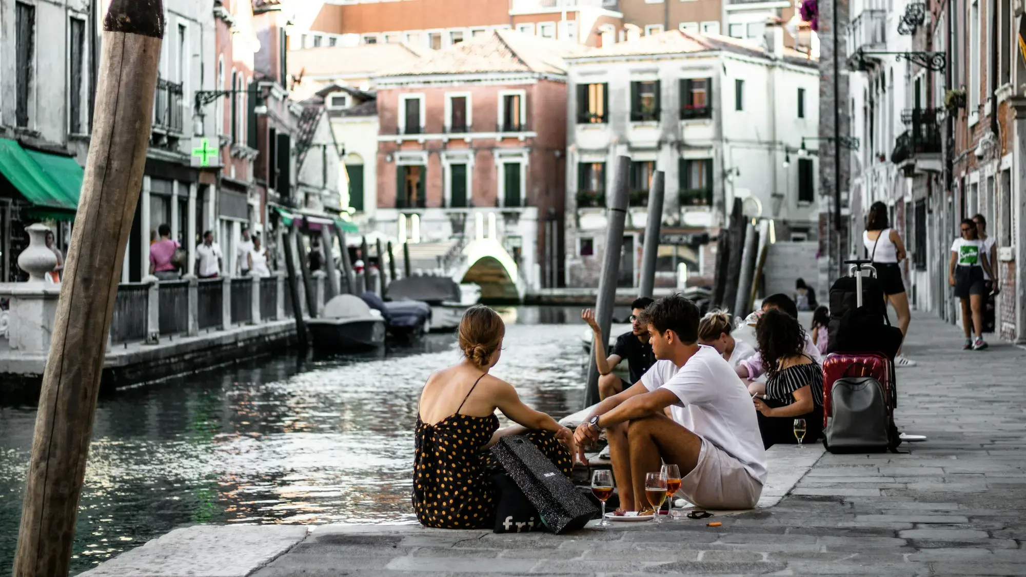Persone sedute lungo un canale veneziano con bicchieri di spritz a inizio serata, con pali d'ormeggio in legno e un piccolo ponte sullo sfondo