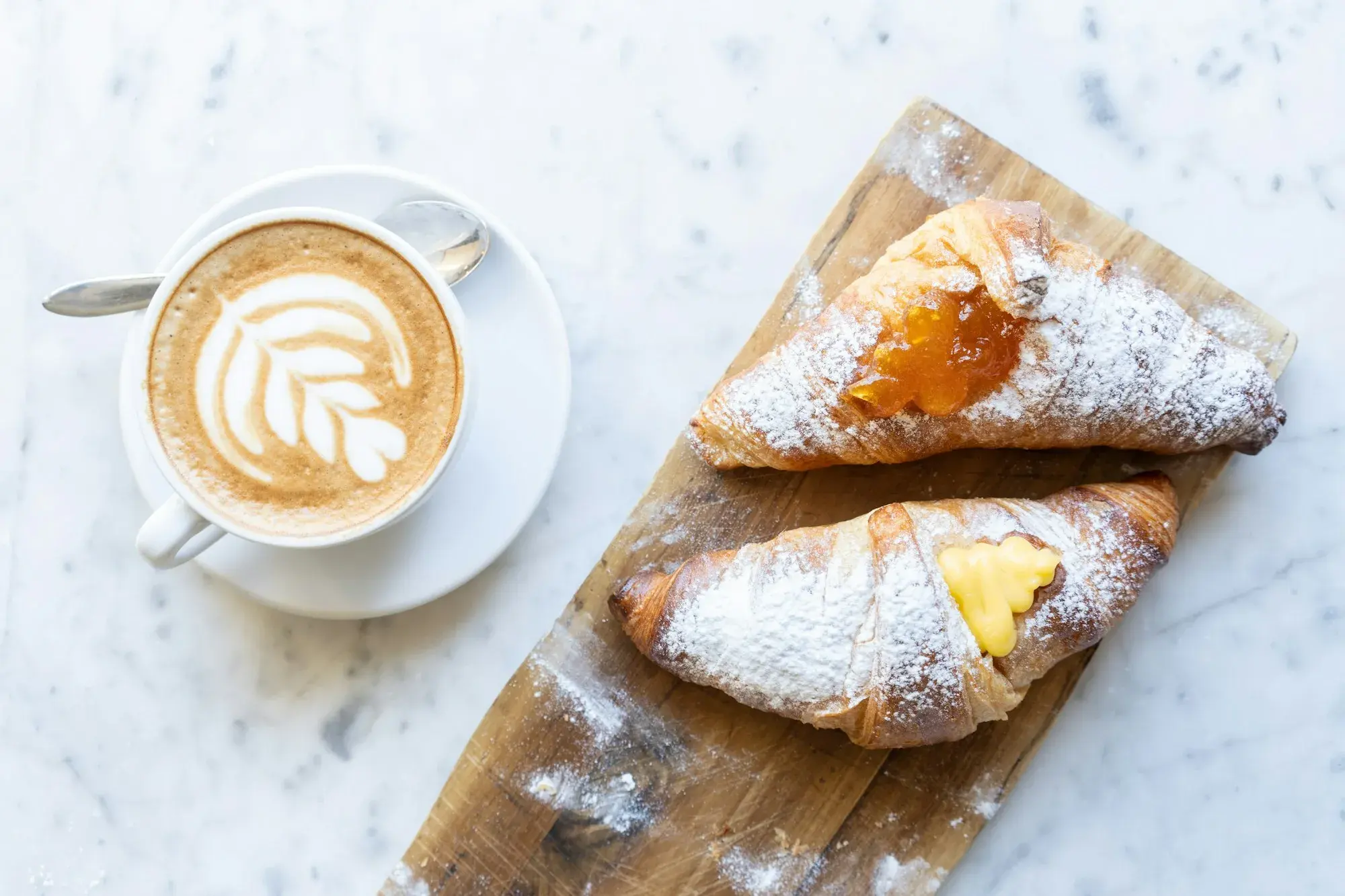 A cappuccino with latte art beside two sugar-dusted cornetti filled with jam and cream on a marble table