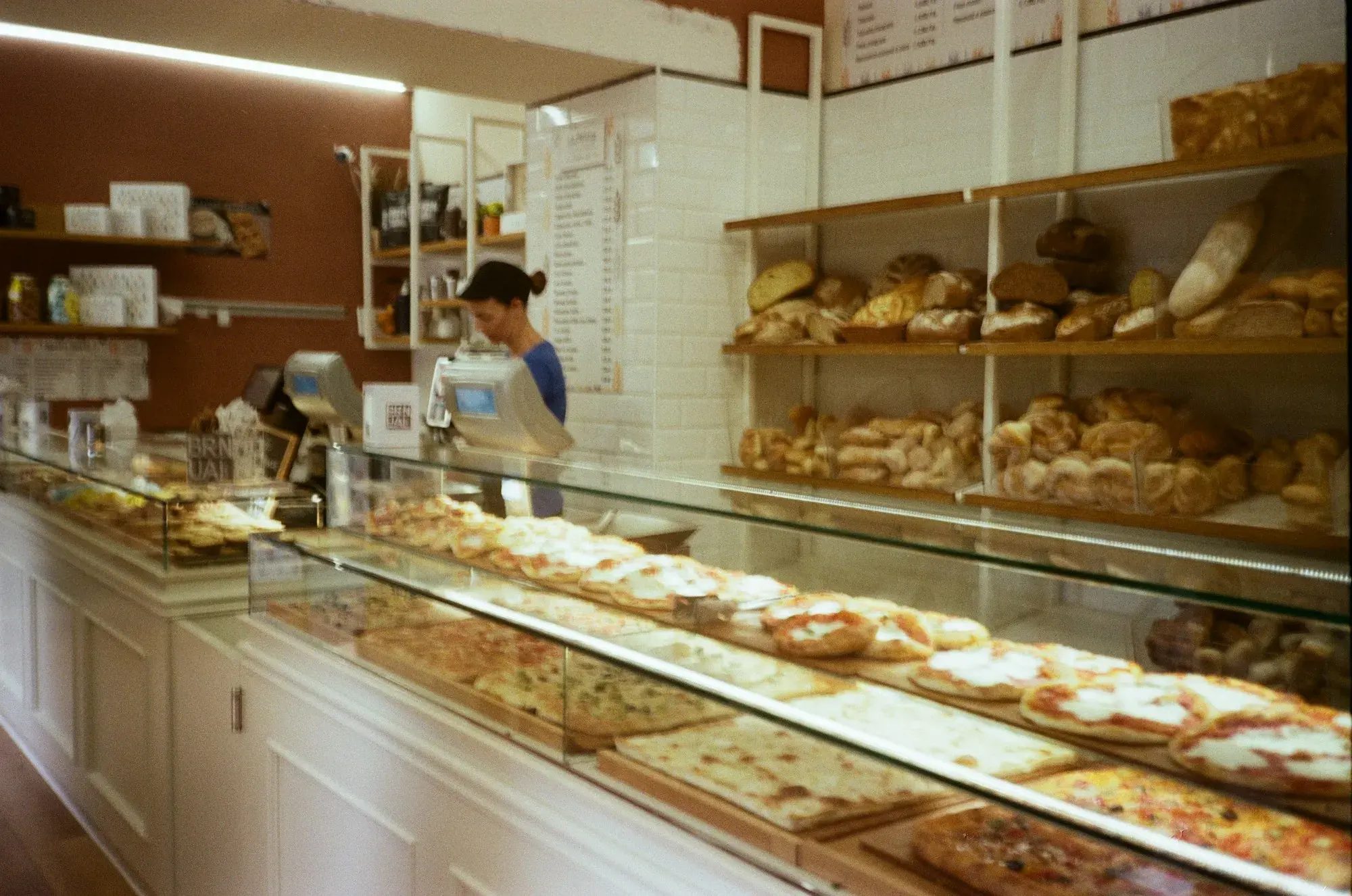 A bakery counter in Genoa with trays of focaccia and loaves on shelves behind, and a baker at work