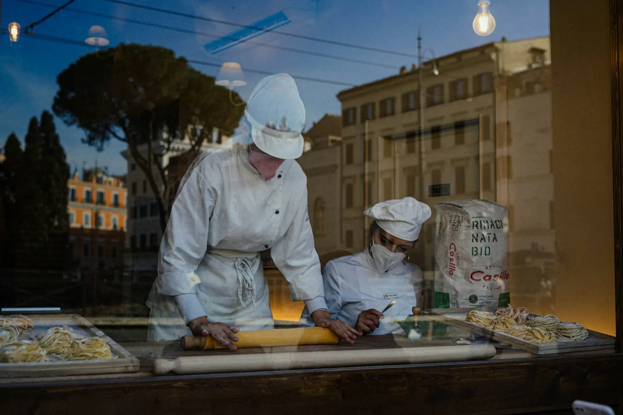 Two cooks in white hats rolling fresh pasta by hand at a counter in a shop window in Rome