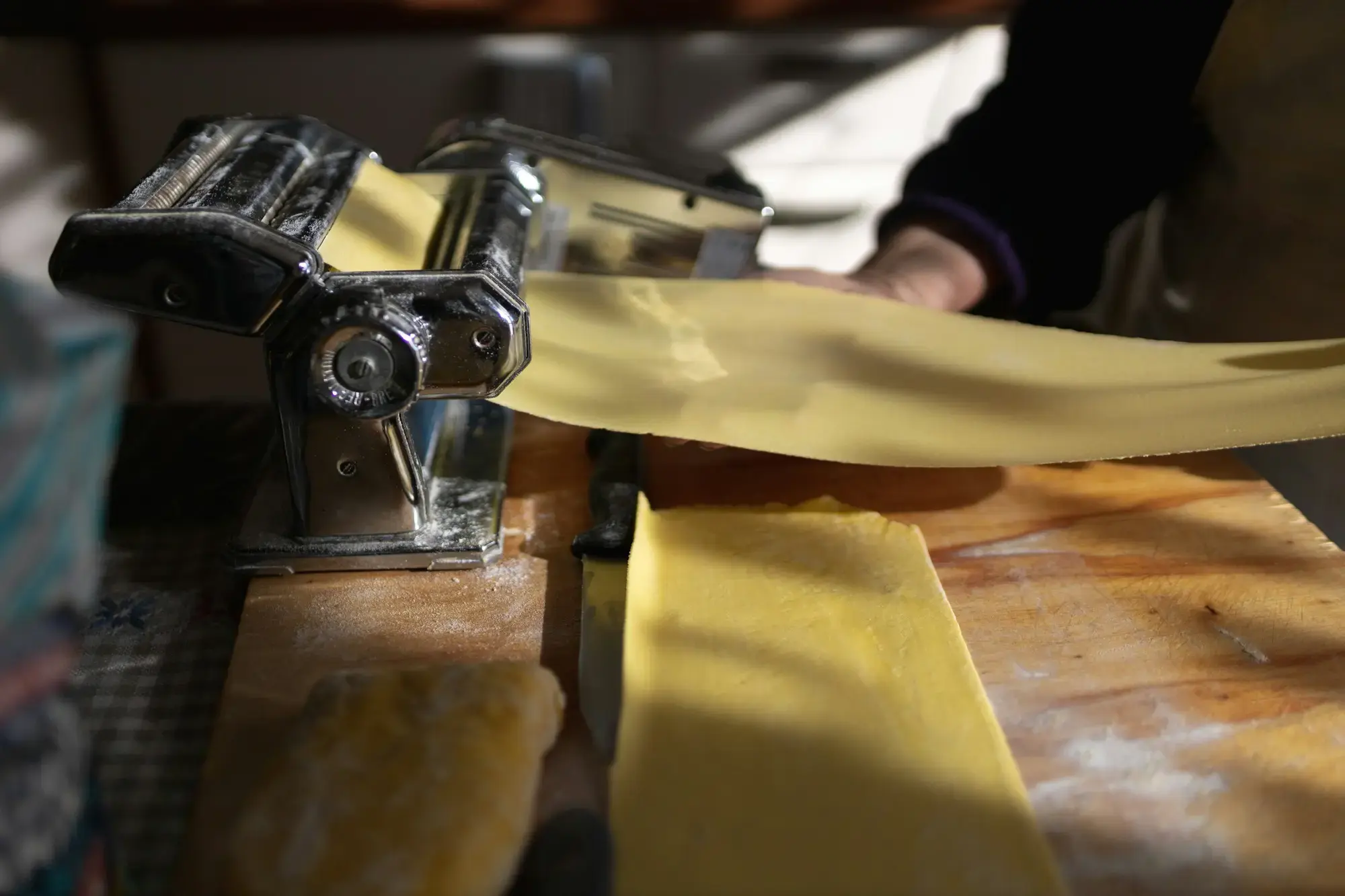 Una persona che passa della pasta fresca attraverso una macchina sfogliatrice, tagliandola in nastri su una superficie infarinata