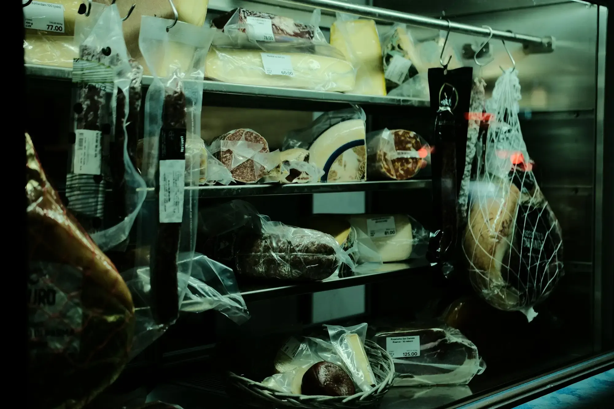An Italian deli display case showing a range of whole and cut cheeses alongside cured meats and olives