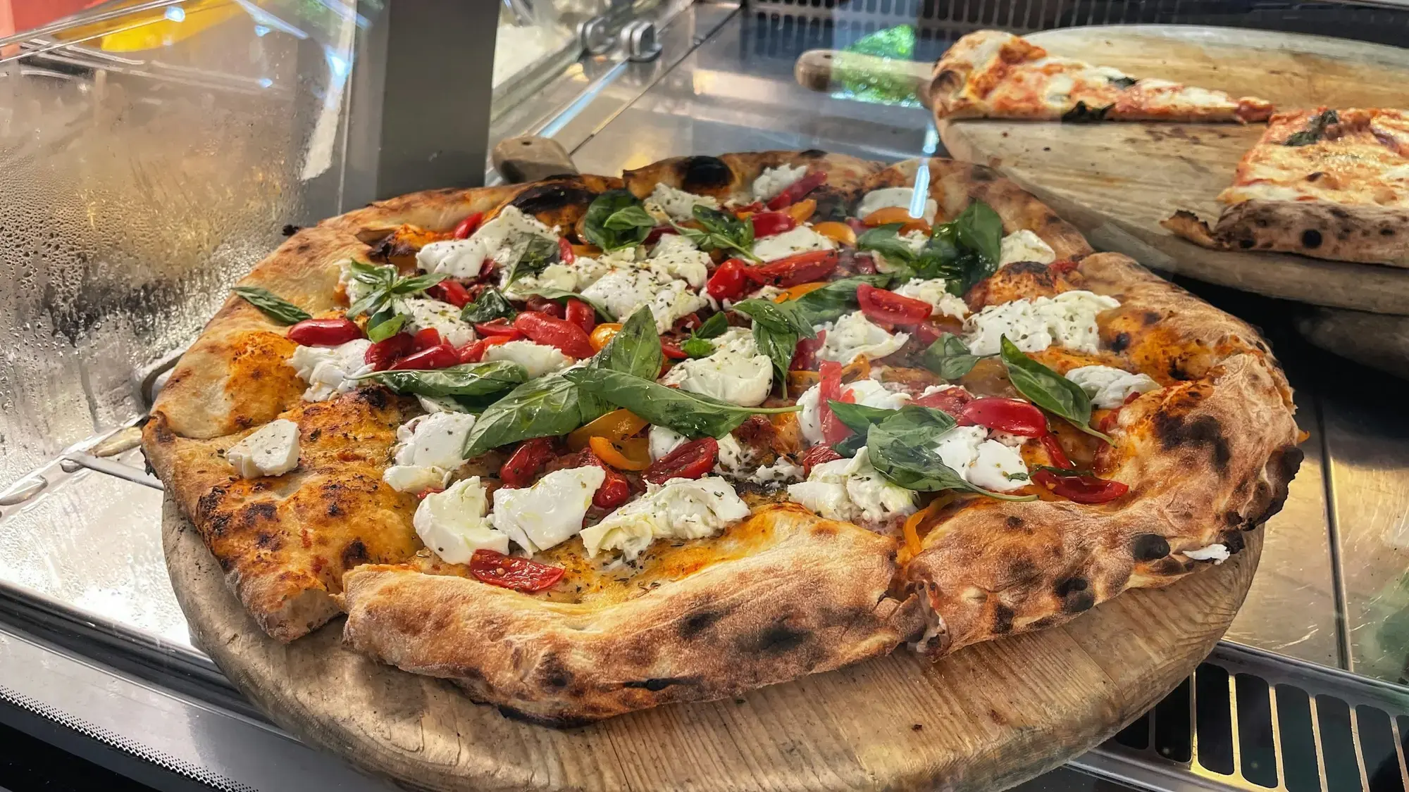A pizza with a puffy, blistered rim topped with torn burrata, cherry tomatoes and basil, on a wooden board at a pizzeria counter in Campania