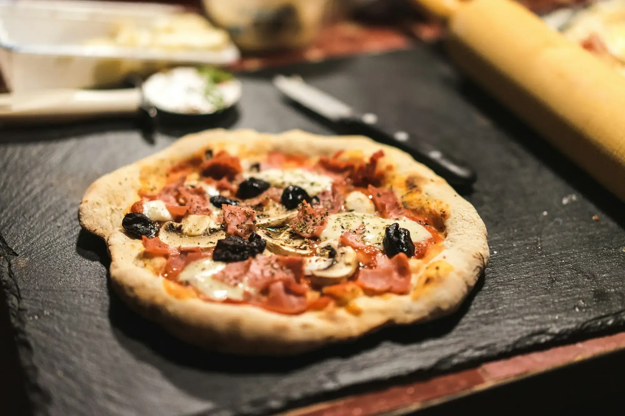 A small homemade pizza with ham, mushrooms and black olives on a slate board, with a rolling pin in the background, made at a home pizza party near Naples