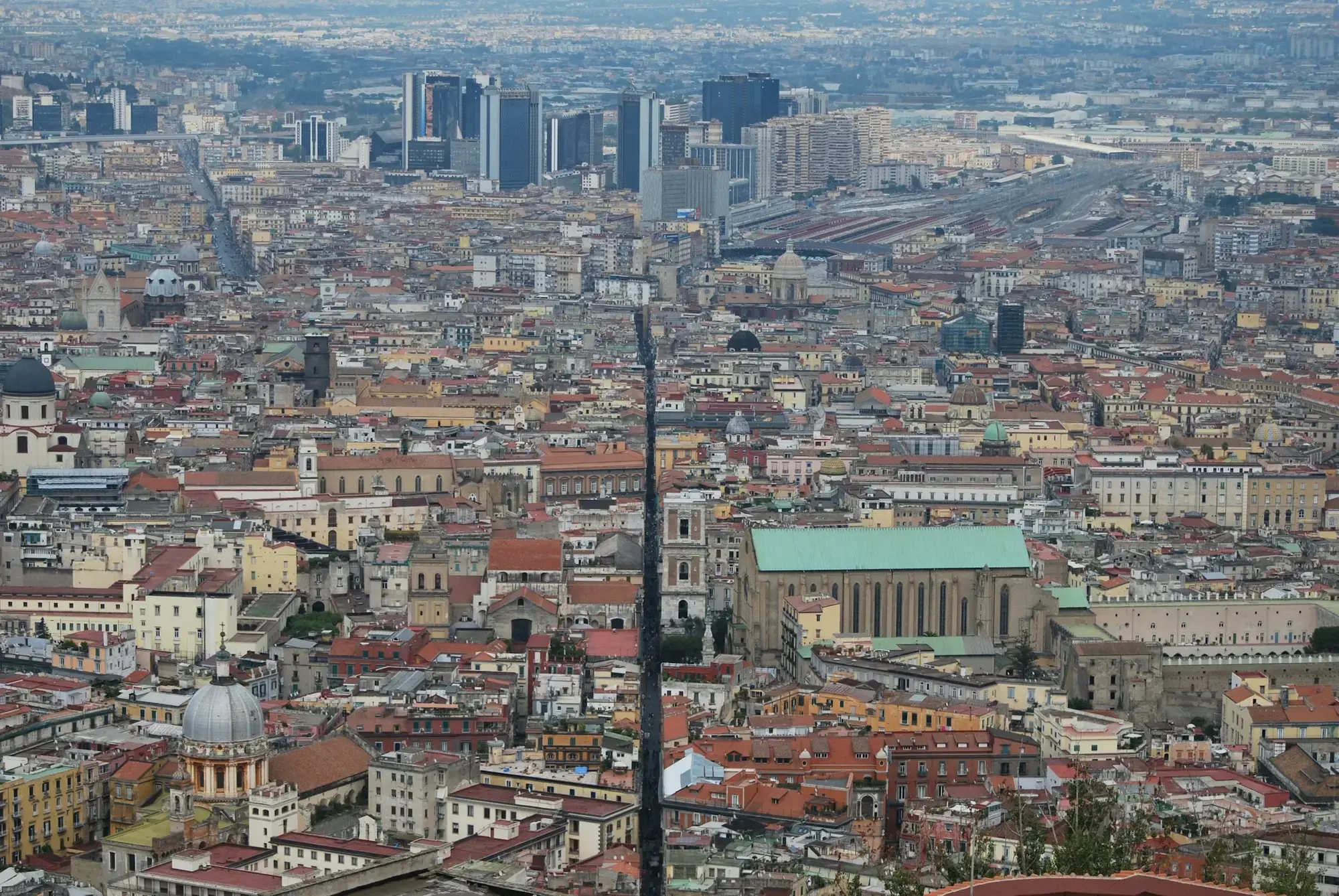 The historic centre of Naples seen from above, a dense grid of rooftops cut through by the long straight street known as Spaccanapoli, with modern towers behind