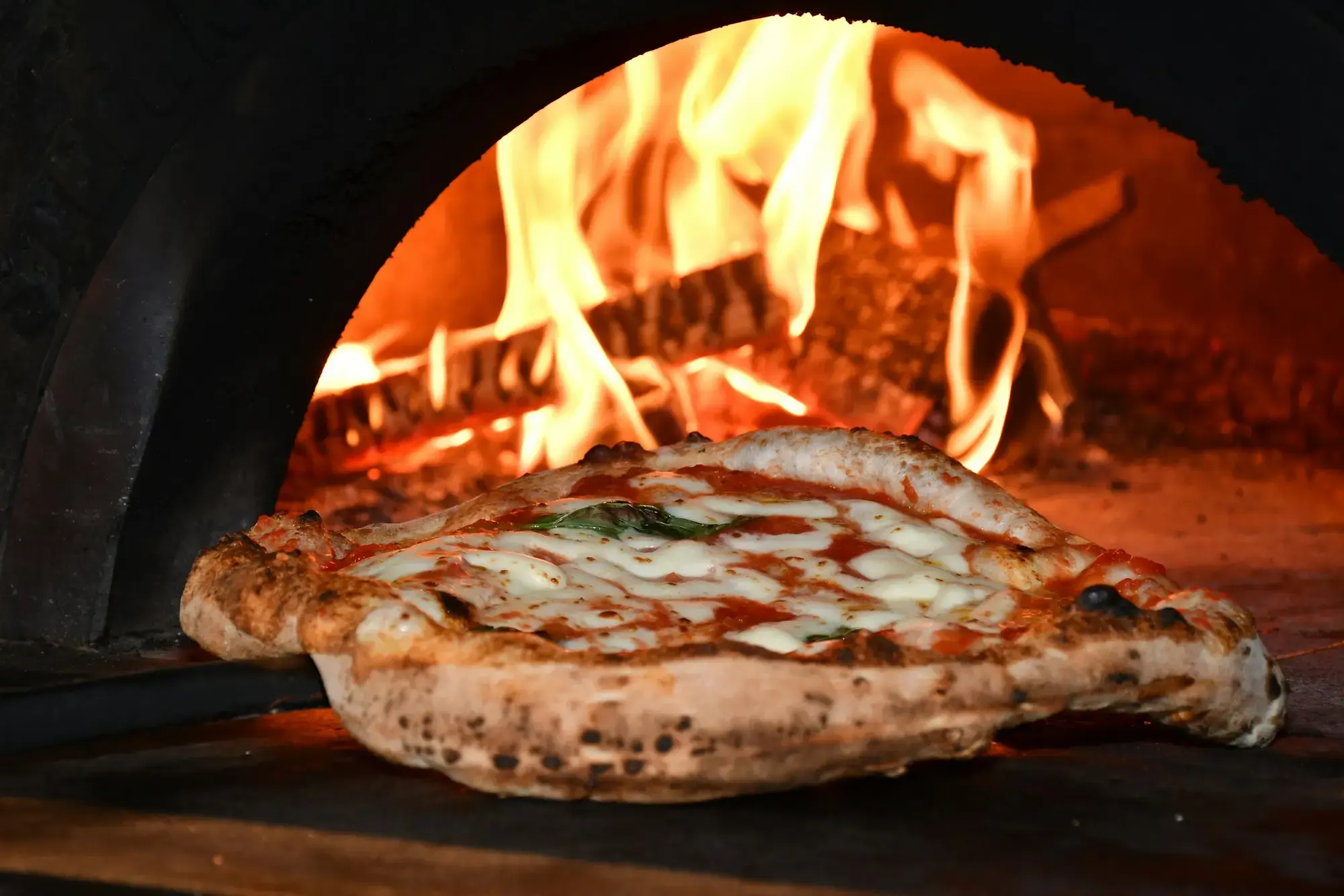 A pizza topped with tomato, melted mozzarella and basil baking on the floor of a wood-fired oven, its rim puffed and blistered, with flames rising behind it
