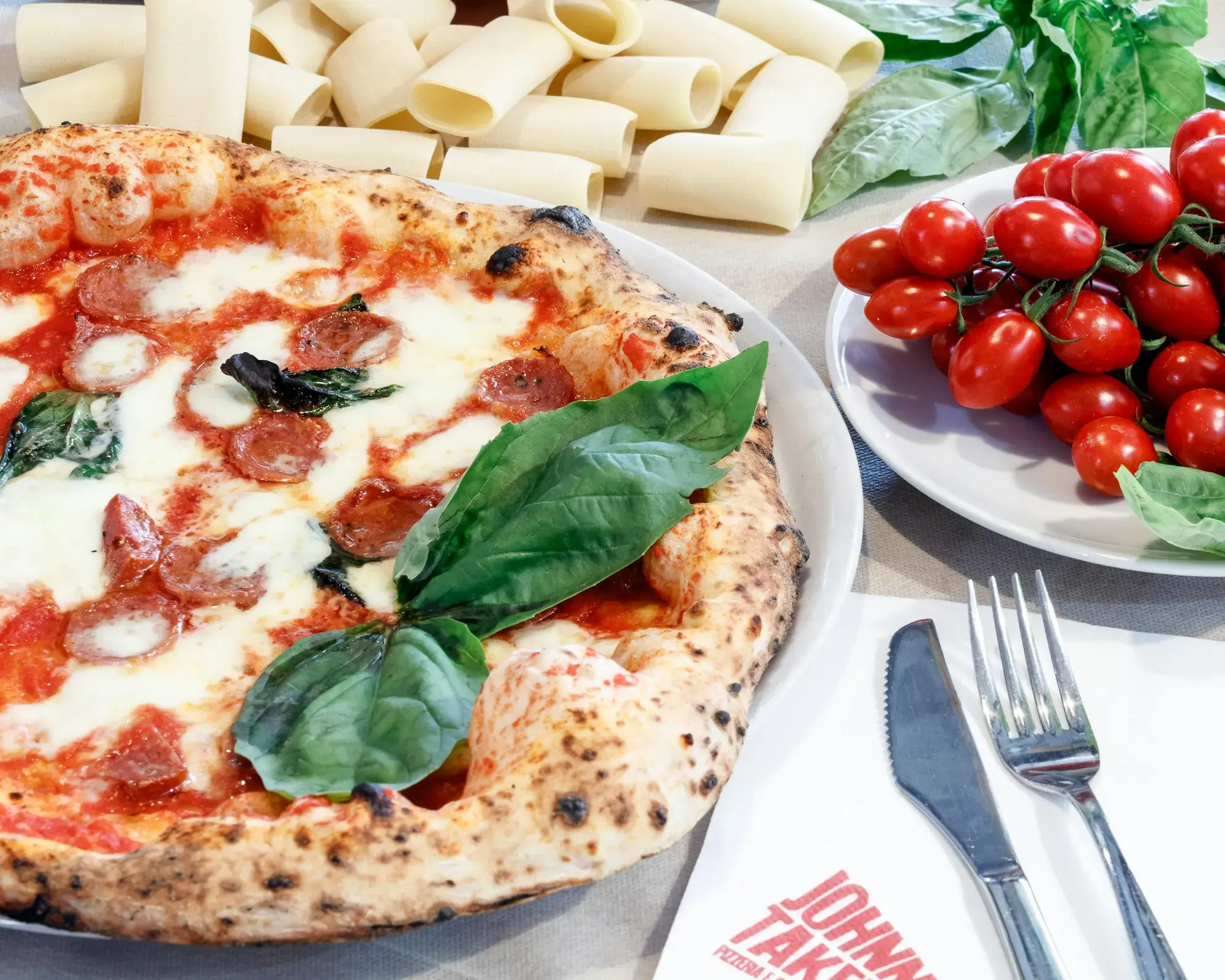 A pizza with tomato, mozzarella, basil and slices of salami served on a plate in Naples, with a knife and fork beside it and cherry tomatoes and pasta in the background
