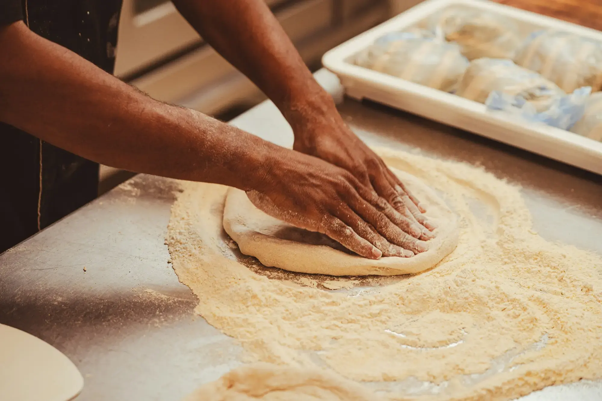 Hands pressing a ball of pizza dough into a disc on a floured marble counter, with trays of dough balls behind