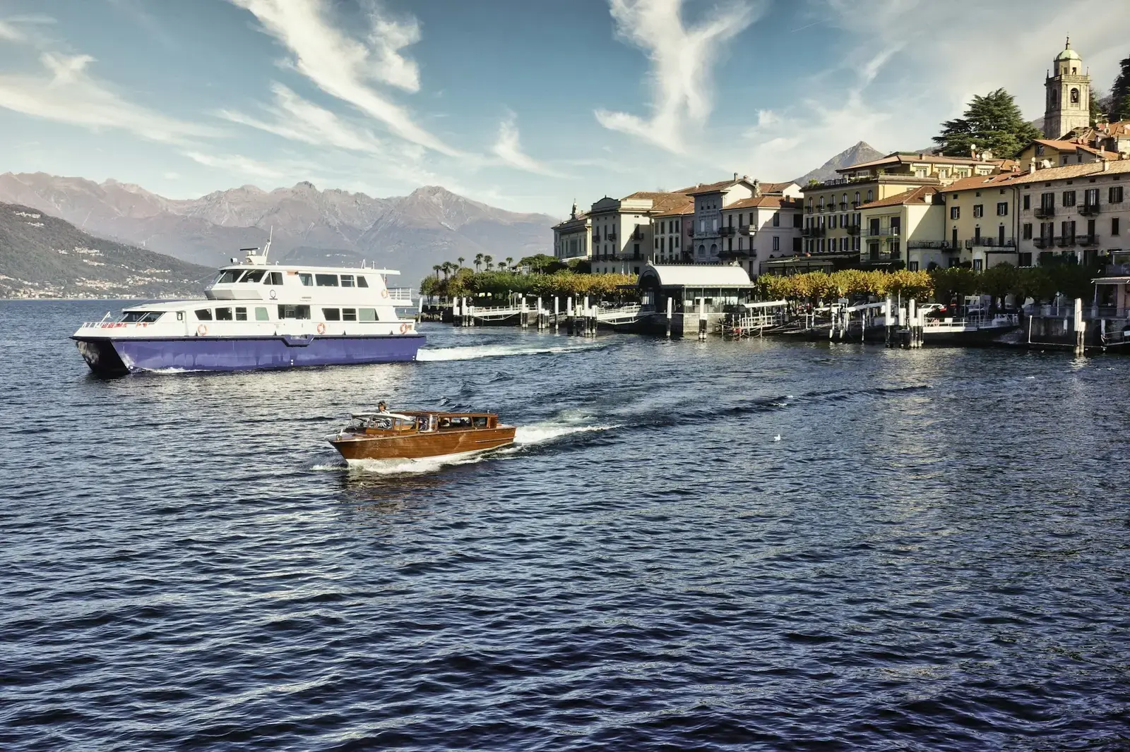A white passenger boat and a wooden motorboat on Lake Como in front of the lakeside town of Bellagio