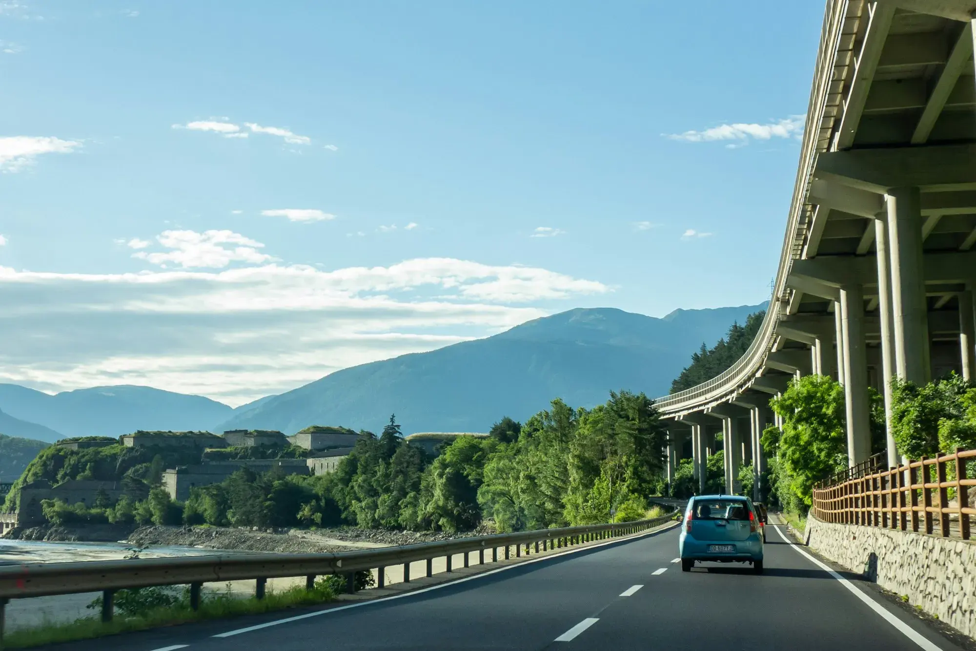 A small car on a road through a green valley in South Tyrol, beside the elevated Brenner motorway viaduct, with mountains behind