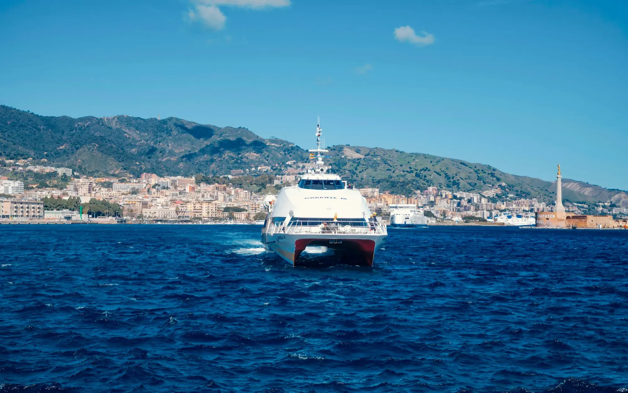 A white fast ferry crossing deep blue water towards the camera, with the city of Messina and hills behind