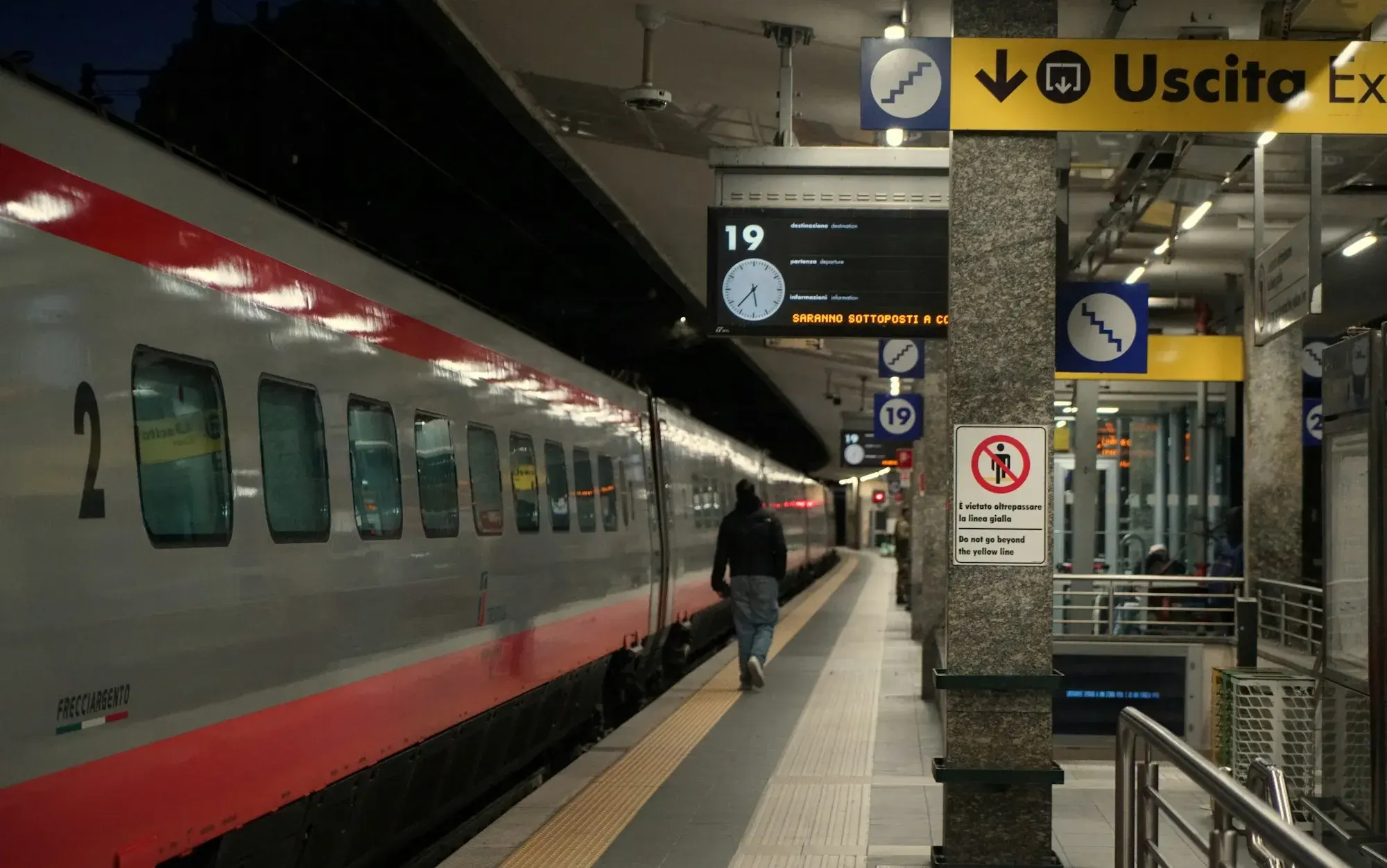 A long-distance train at a platform at Pisa station, with a digital departure screen, clock and yellow Uscita exit sign